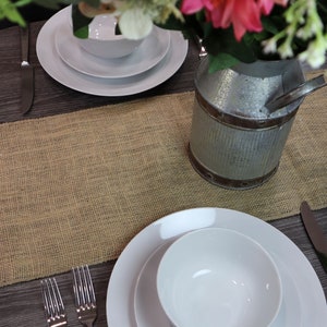 May include: A rustic brown burlap table runner with a white ceramic bowl and plate on a dark wood table. A galvanized metal pitcher with a bouquet of pink and white flowers sits in the center of the runner.