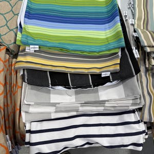 May include: A stack of outdoor cushions in various patterns and colors. The cushions feature stripes in blue, green, yellow, and gray, as well as black and white checkered and striped designs. Some cushions have tags attached.