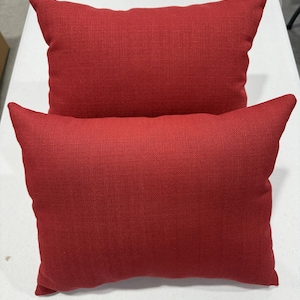 May include: Two rectangular red throw pillows with pointed corners. The pillows have a textured fabric and are arranged one on top of the other. The pillows are a solid, vibrant red color.