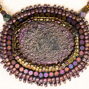 Può includere: Una collana fatta a mano con un pendente ovale con un centro scuro e strutturato. Il pendente è circondato da file di piccole perline iridescenti nei toni del viola, marrone e oro. La collana ha una catena di perline.