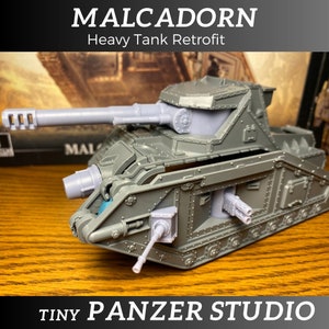 May include: A gray heavy tank retrofit model with the text "MALCADORN Heavy Tank Retrofit" and "TINY PANZER STUDIO". The model features a long gun barrel and multiple smaller cannons, showcasing detailed design elements.