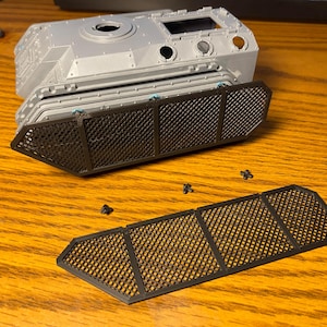 May include: A gray plastic model tank body with a black mesh panel and separate black mesh pieces. Small black screws are scattered on the wooden surface. The tank has various openings and details.