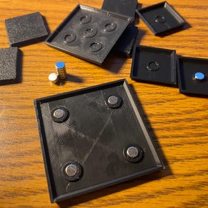 May include: Black square trays with embedded magnets and small cylindrical magnets are scattered on a wooden surface. Some trays have circular indentations. Two textured black squares are also present.
