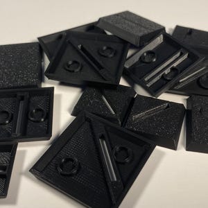 May include: A collection of black 3D-printed geometric shapes, including squares, triangles, and rectangles. Some pieces have circular indentations and linear grooves. The objects are scattered on a white surface.