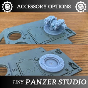 Puede incluir: Se muestran dos accesorios grises para tanques en miniatura. Uno tiene una torreta con tres cañones, el otro una escotilla circular. Los textos "ACCESSORY OPTIONS" y "TINY PANZER STUDIO" son visibles.