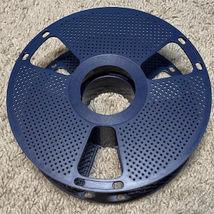 May include: A dark blue spool of filament for 3D printing. The spool has a circular design with numerous small holes and several larger openings around the perimeter. The center has a large circular opening.