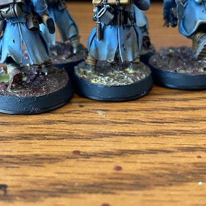May include: Miniature figurines of soldiers in blue coats and detailed gear, standing on textured, circular bases. The bases are painted with earth-tone colors, suggesting a battlefield setting. The figures are positioned on a wooden surface.