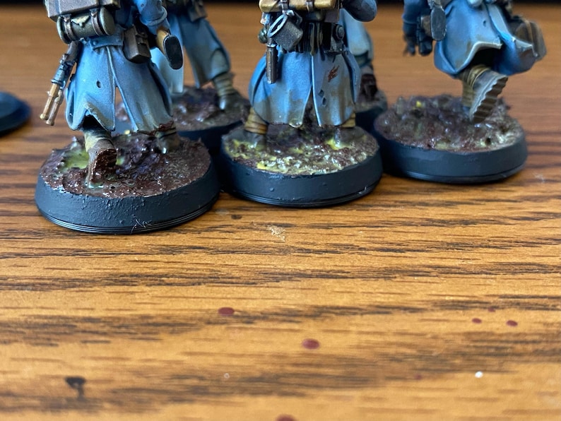 May include: Miniature figurines of soldiers in blue coats and detailed gear, standing on textured, circular bases. The bases are painted with earth-tone colors, suggesting a battlefield setting. The figures are positioned on a wooden surface.