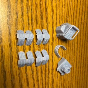 Puede incluir: Un conjunto de componentes impresos en 3D de color gris claro, que incluyen varias piezas segmentadas, una peque&ntilde;a estructura en forma de caja con un asa y una pieza curva. Las piezas est&aacute;n dispuestas sobre una superficie de madera.
