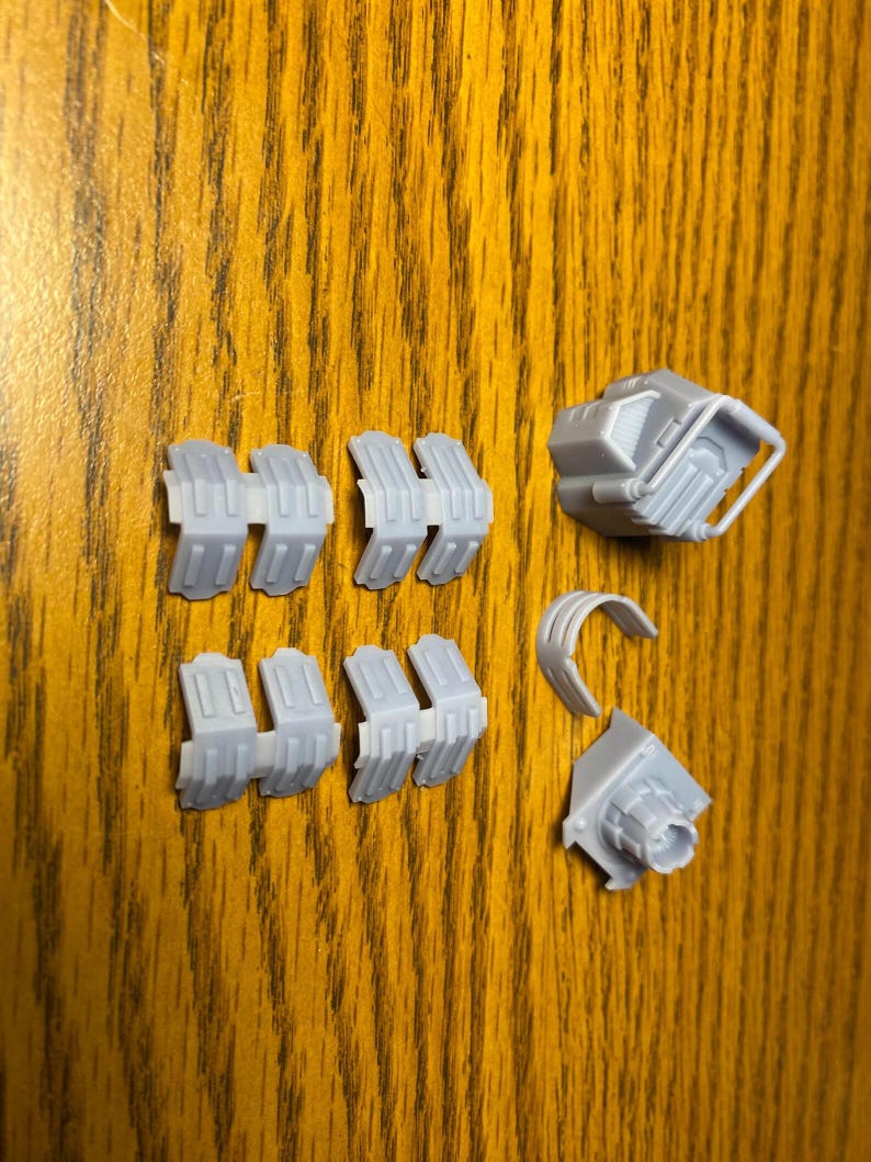 Puede incluir: Un conjunto de componentes impresos en 3D de color gris claro, que incluyen varias piezas segmentadas, una peque&ntilde;a estructura en forma de caja con un asa y una pieza curva. Las piezas est&aacute;n dispuestas sobre una superficie de madera.