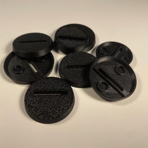 May include: A collection of black, circular bases with textured surfaces. Each base has a slot or indentation, designed for miniature figures. The bases are likely for tabletop gaming or model building.