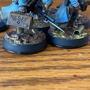 May include: Miniature figurines of soldiers, painted in blue and brown, with detailed weaponry and accessories. Each figure is mounted on a circular base with textured terrain. The figures are set against a wooden surface.