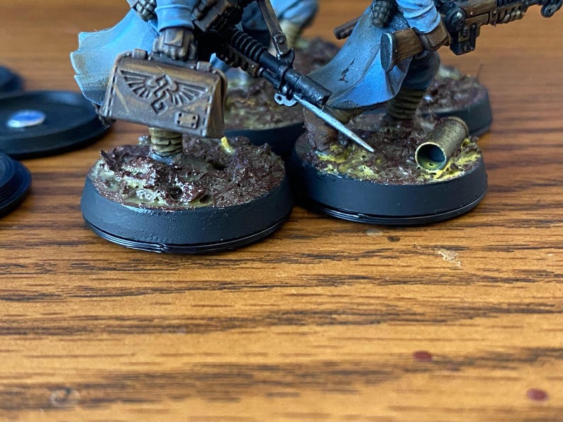 May include: Miniature figurines of soldiers, painted in blue and brown, with detailed weaponry and accessories. Each figure is mounted on a circular base with textured terrain. The figures are set against a wooden surface.