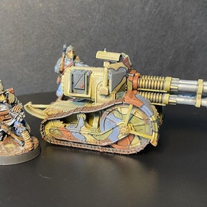 Puede incluir: Un modelo de juego de guerra en miniatura de un vehículo de orugas con dos cañones grandes. El vehículo está pintado con un patrón de camuflaje marrón, verde y azul. Un soldado en miniatura está de pie junto al vehículo.