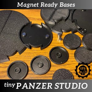 May include: Black plastic magnet-ready bases in various shapes and sizes, including round and segmented designs. The bases have textured surfaces and embedded magnets. The text "Magnet Ready Bases" and "tiny PANZER STUDIO" are visible.