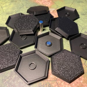 Op de afbeelding: Een verzameling zwarte, zeshoekige spelstukken met een getextureerd en glad oppervlak. Sommige stukken hebben een centrale ring of een magneet met een blauwe bovenkant. De stukken liggen op een groen en beige spelbord met zeshoekige rasterlijnen.
