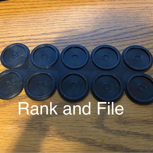 May include: A black plastic component with ten circular pieces connected in a row. The text "Rank and File" is printed in white below the component. The items are on a wooden surface.
