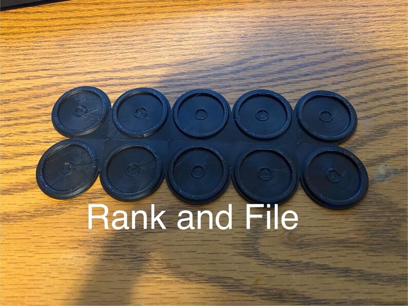 May include: A black plastic component with ten circular pieces connected in a row. The text "Rank and File" is printed in white below the component. The items are on a wooden surface.