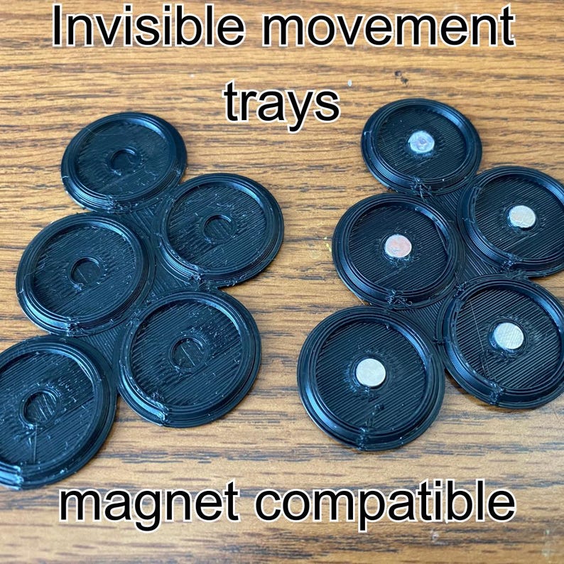 May include: Black, circular trays with a central hole and a textured surface. The trays are arranged in clusters of six, with some containing small, silver magnets. The text "Invisible movement trays" and "magnet compatible" is visible.
