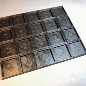 May include: A black plastic tray with 25 square compartments, each with a circular cutout in the centre.