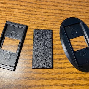 May include: Three black plastic 3D printed shapes. The first shape is a rectangle with a rectangular cutout in the centre. The second shape is a rectangle with a smooth surface. The third shape is an oval with a rectangular cutout in the centre.