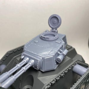 May include: A gray model tank turret with two gun barrels and an open hatch. The turret is mounted on a dark green tank body. The model appears to be made of plastic and is designed for hobbyists or collectors.