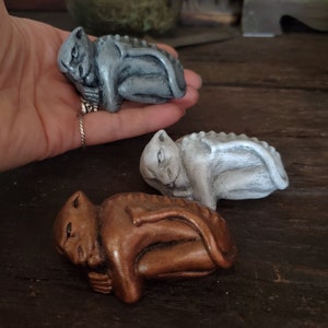 May include: Three small gargoyle figurines in gray, white, and bronze. The gargoyles are in a resting pose, with their wings folded and heads resting on their paws. The figurines are on a dark wooden surface.