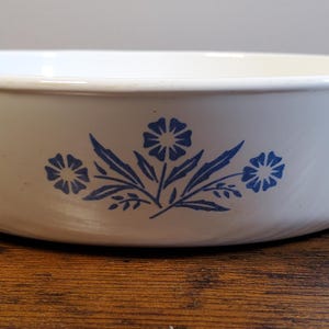 May include: A white ceramic bowl with a blue floral design. The design features stylized flowers and leaves. The bowl is set on a wooden surface. The bowl's rim is visible.