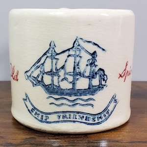 May include: A small, off-white ceramic mug with a blue illustration of a sailing ship. The words "Old" and "Spirits" are in red script on either side of the ship. Below the ship is a banner that reads "Ship Friendship" in blue.