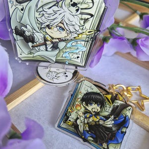 Könnte beinhalten: Zwei Acryl-Aufsteller mit Anime-Illustrationen. Einer zeigt eine weißhaarige Figur mit Monokel und Federkiel über einem offenen Buch. Der andere zeigt eine dunkelhaarige Figur mit Ziegenbart, blauem Umhang und Flamme über einem Buch.
