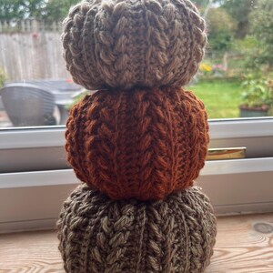 May include: A stack of three crocheted pumpkins featuring a textured cable knit pattern. The top pumpkin is light brown, the middle pumpkin is a deep burnt orange, and the bottom pumpkin is a muted olive green. Each pumpkin has a small brown stem.