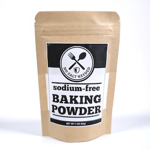 May include: A brown paper bag of sodium-free baking powder. The label features a black and white design with a fork and spoon crossed over a shield, and the words "No Salt Needed". The text "sodium-free BAKING POWDER" is printed in bold, black letters. The bag contains 3 oz (85g) of product.