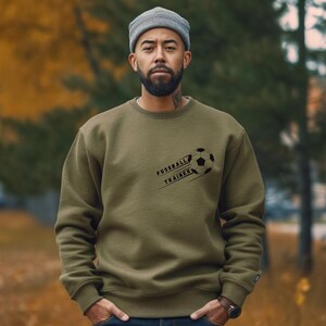 Könnte beinhalten: Olivgrünes Sweatshirt mit dem Text "FUSSBALL TRAINER" und einer Fußballgrafik. Die Person trägt eine graue Mütze.
