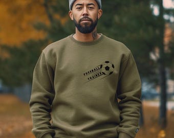 Op de afbeelding: Olijfgroene sweatshirt met de tekst "FUSSBALL TRAINER" en een voetbalafbeelding. De persoon draagt een grijze muts.