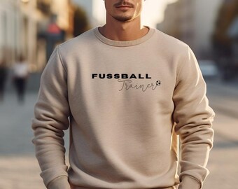 Op de afbeelding: Beige crewneck sweatshirt met de tekst "FUSSBALL Trainer" in zwarte letters. Het woord "Trainer" is in een cursief lettertype geschreven, met een voetbalafbeelding. De sweatshirt heeft lange mouwen en een geribbelde halslijn.