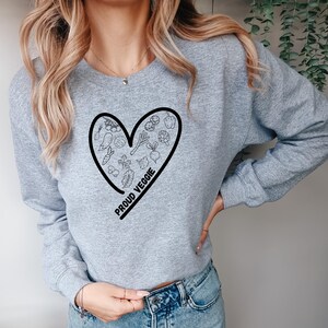 Könnte beinhalten: Hellgraues Sweatshirt mit einer schwarzen herzförmigen Grafik, die mit Gemüseillustrationen gefüllt ist. Die Worte "PROUD VEGGIE" sind in Schwarz auf der unteren linken Seite des Herzens aufgedruckt.