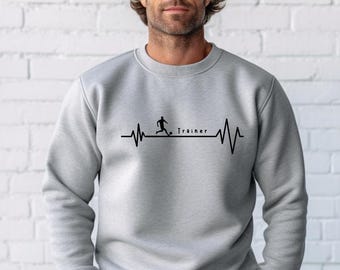 Op de afbeelding: Een lichtgrijs sweatshirt met een zwarte afbeelding van een voetballer die een bal trapt, geïntegreerd in een hartslaglijnontwerp. Het woord "Trainer" is zwart gedrukt. Het sweatshirt is gemaakt van zacht materiaal.