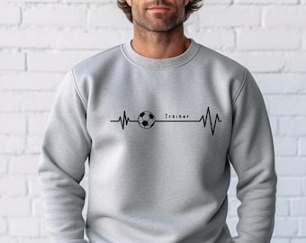 Op de afbeelding: Een lichtgrijs sweatshirt met een voetbalontwerp geïntegreerd in een hartslaglijn. Het woord "Trainer" is in het zwart gedrukt. De sweatshirt heeft een ronde hals.