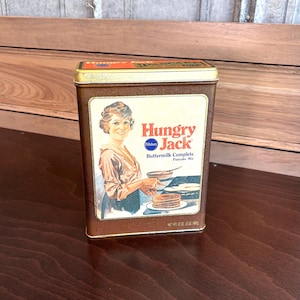 May include: A vintage tin box for Hungry Jack Buttermilk Complete Pancake Mix. The box features a woman in a yellow dress making pancakes in a pan. The box is brown with gold trim and the text "Hungry Jack" in red and white.