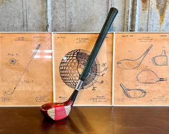 May include: A vintage golf club with a red and white grip and a green and black shaft. The club is resting on a wooden surface in front of three framed patent drawings for golf balls and clubs.