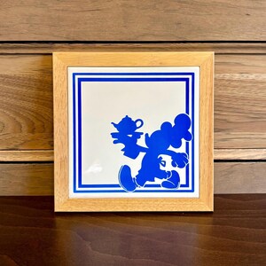 May include: A framed blue and white tile with a silhouette of a cartoon character holding a teapot. The character is walking and has a large head and small body.