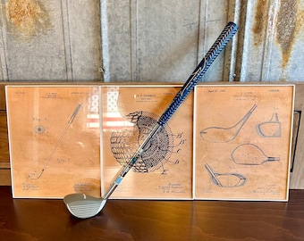 May include: A vintage golf club with a blue and white grip, resting on a wooden surface in front of three framed vintage golf patent drawings. The drawings are in black and white and feature illustrations of golf clubs and golf balls.