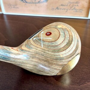 Puede incluir: Primer plano de una cabeza de palo de golf de madera vintage con un patr&oacute;n de vetas de madera marr&oacute;n claro y verde. La cabeza del palo tiene un dise&ntilde;o de cruz blanca y un punto rojo. Una pieza de metal dorado est&aacute; en la parte inferior.