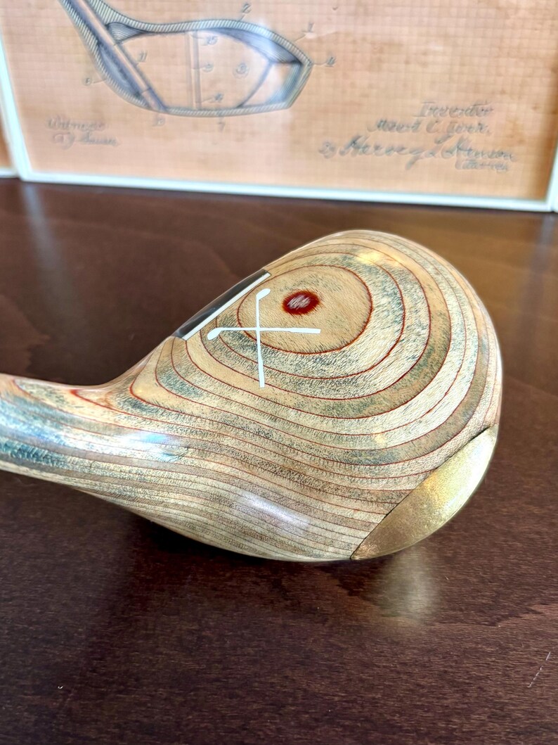 Puede incluir: Primer plano de una cabeza de palo de golf de madera vintage con un patr&oacute;n de vetas de madera marr&oacute;n claro y verde. La cabeza del palo tiene un dise&ntilde;o de cruz blanca y un punto rojo. Una pieza de metal dorado est&aacute; en la parte inferior.
