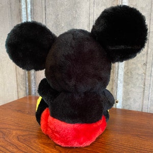 May include: A plush Mickey Mouse toy, viewed from the back, with large black ears. The toy has a black body, red shorts, and yellow accents. The toy is sitting on a wooden surface.