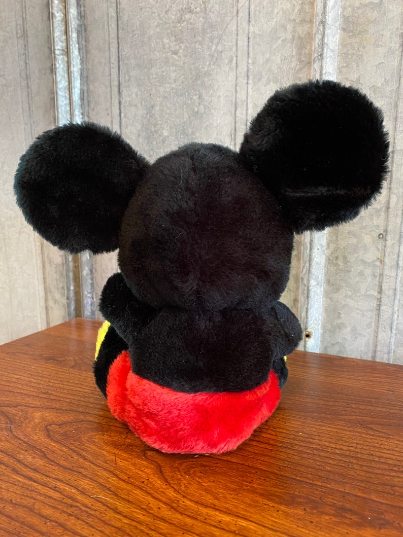 May include: A plush Mickey Mouse toy, viewed from the back, with large black ears. The toy has a black body, red shorts, and yellow accents. The toy is sitting on a wooden surface.