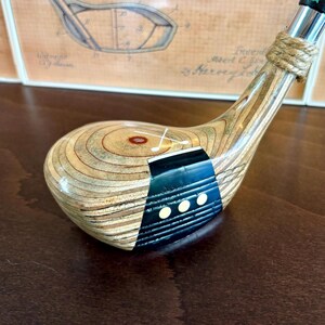 Puede incluir: Un palo de golf de madera vintage con una cabeza marr&oacute;n claro y un acento azul oscuro y negro. El palo presenta un cuello envuelto en cuerda y un agarre verde y negro. El fondo incluye una ilustraci&oacute;n de patente enmarcada.