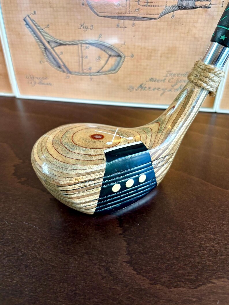 Puede incluir: Un palo de golf de madera vintage con una cabeza marr&oacute;n claro y un acento azul oscuro y negro. El palo presenta un cuello envuelto en cuerda y un agarre verde y negro. El fondo incluye una ilustraci&oacute;n de patente enmarcada.