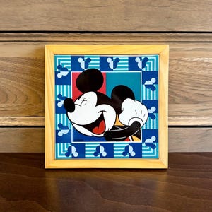 May include: A framed picture of Mickey Mouse with a blue, green, and red background. The background is a pattern of Mickey Mouse ears. The picture is in a light wood frame.