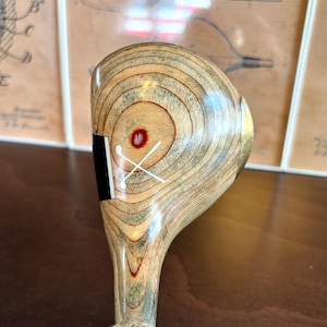 Puede incluir: Un palo de golf de madera vintage con un patr&oacute;n de vetas de madera marr&oacute;n claro y beige. La cabeza del palo presenta un acento negro y un dise&ntilde;o de tee de golf cruzado blanco. El agarre est&aacute; envuelto con hilo natural.